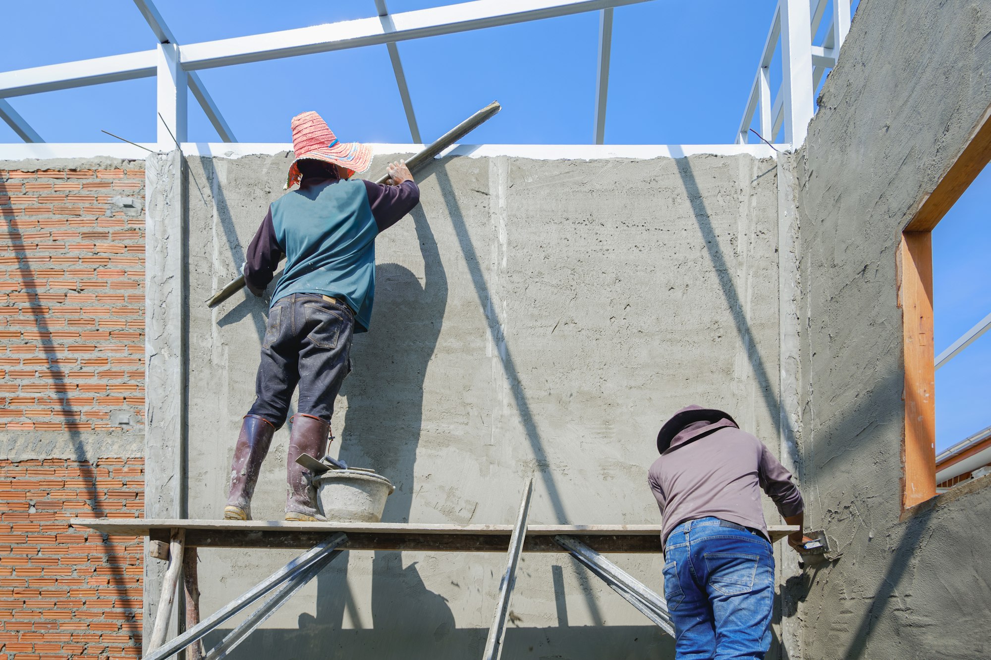 2 Asian builder workers on scaffolding are plastering concrete wall room in house construction site